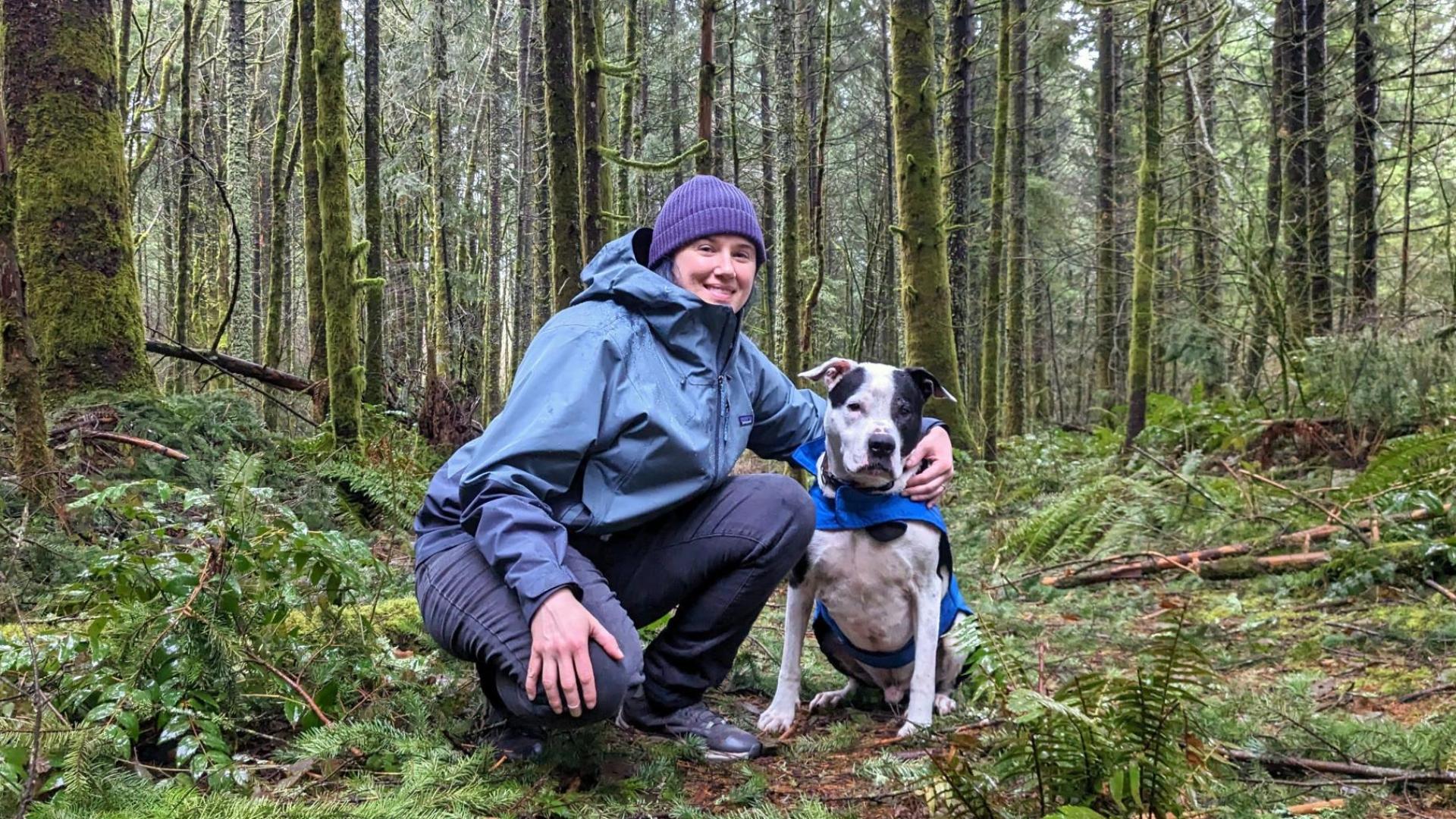 Project Drawdown scientist in a green raincoat crouches with her arm around a dog in a blue raincoat among ferns and moss-covered trees.