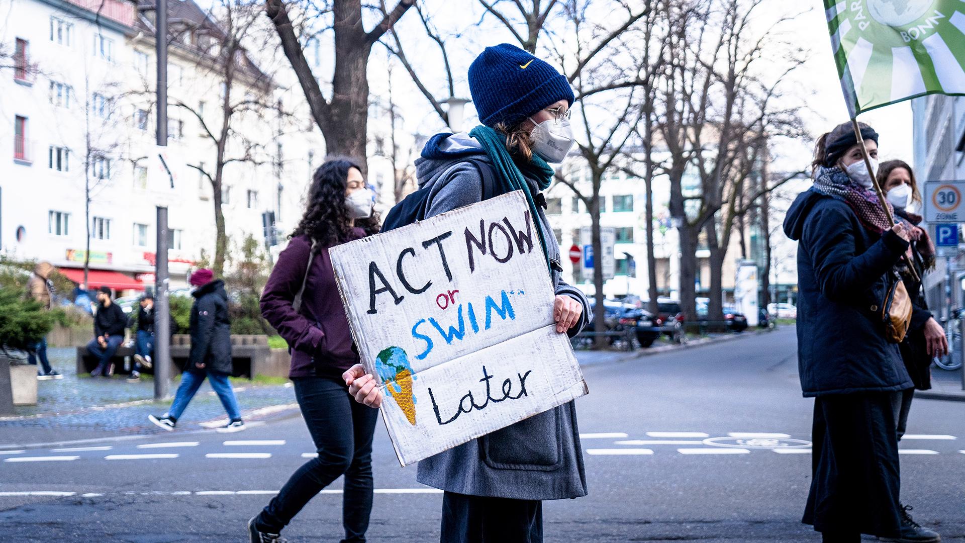 A woman holding a sign that reads "Act Now or Swim Later"